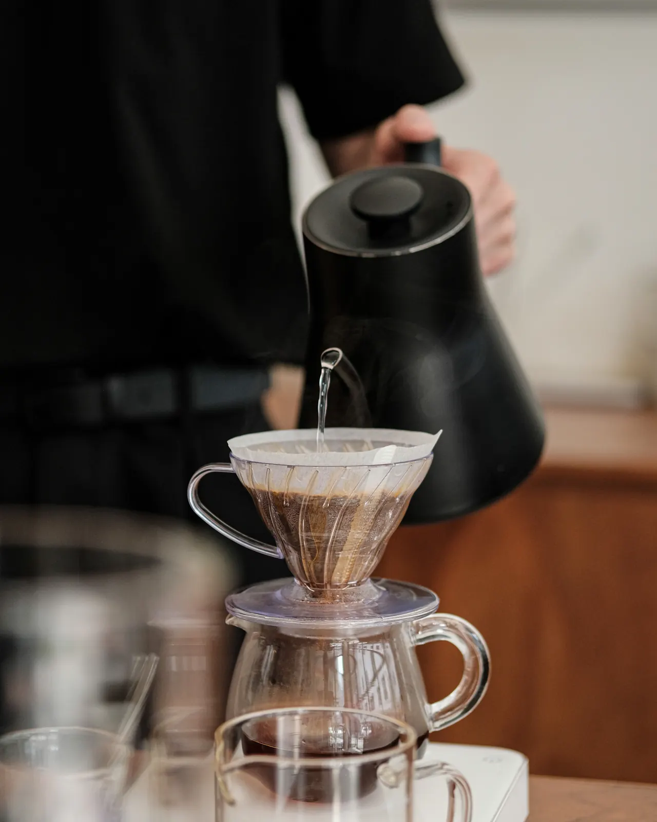 Barista menuang air panas dari ketel hitam ke dripper V60 di atas meja kedai